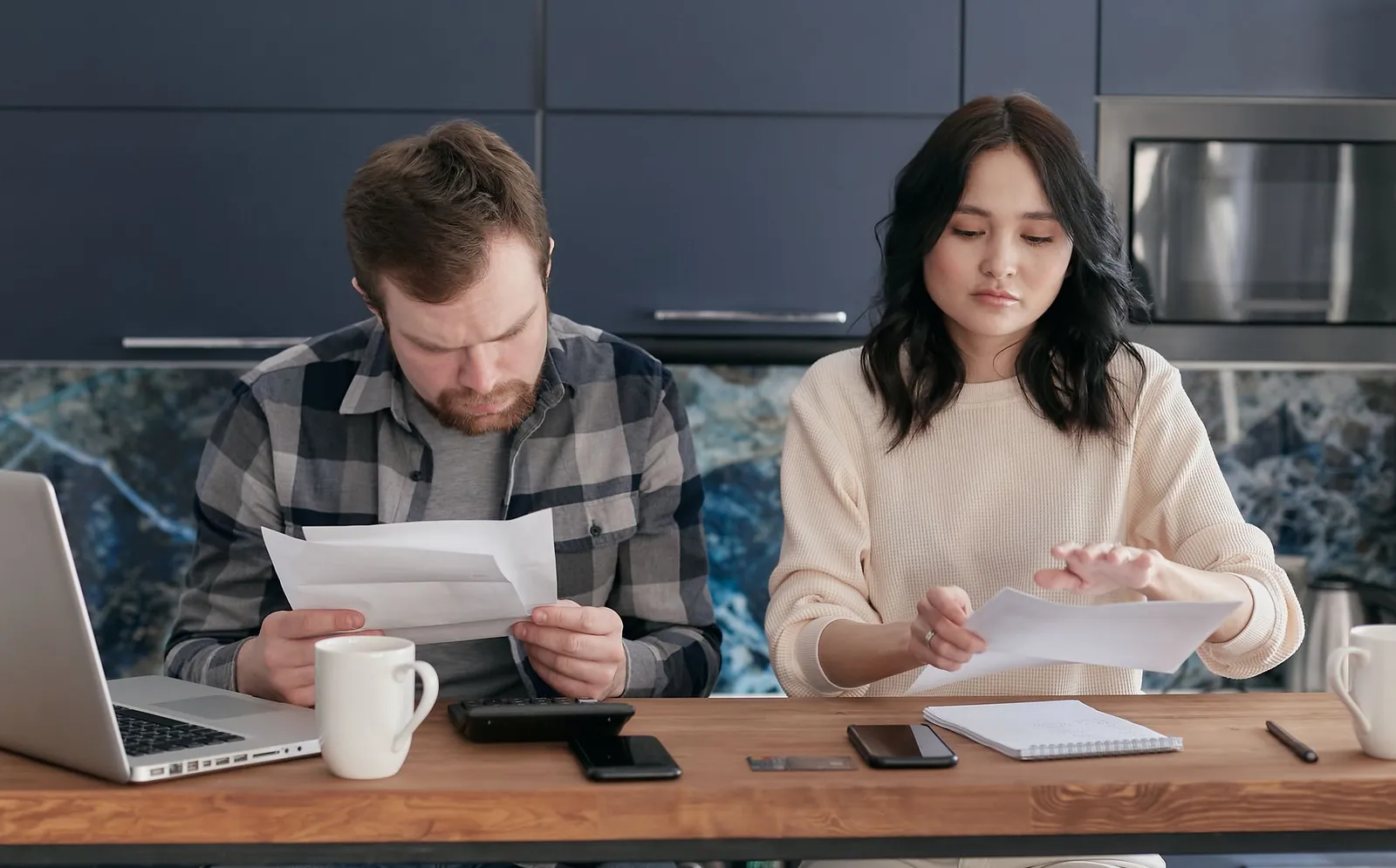 Couple reading bills at a kitchen counter with a calculator, phones and a laptop
