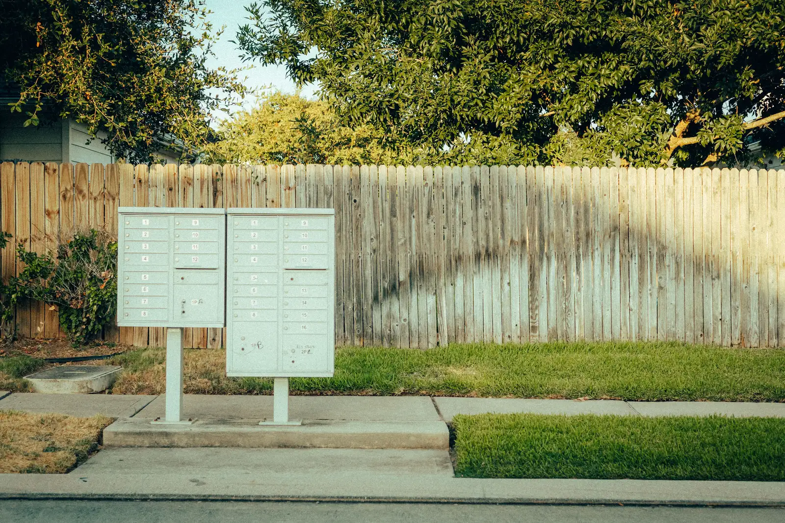 Two cluster mailboxes on a concrete pad in front of a wooden fence on a sunny suburban street