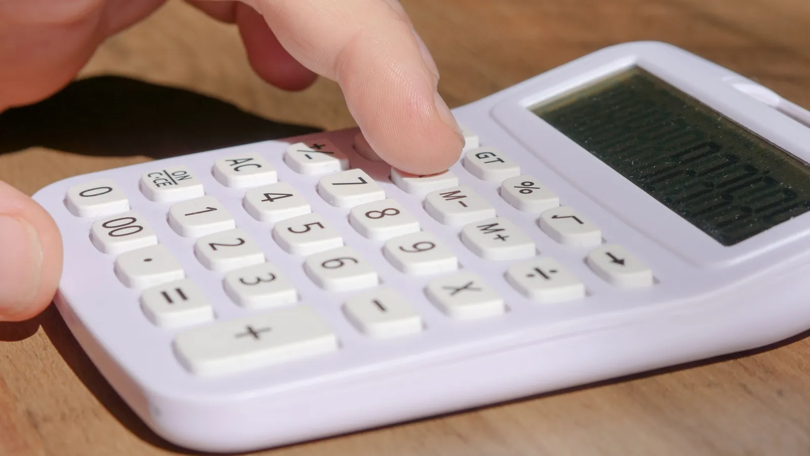 A finger pressing the keys of a white desktop calculator on a sunlit wooden table