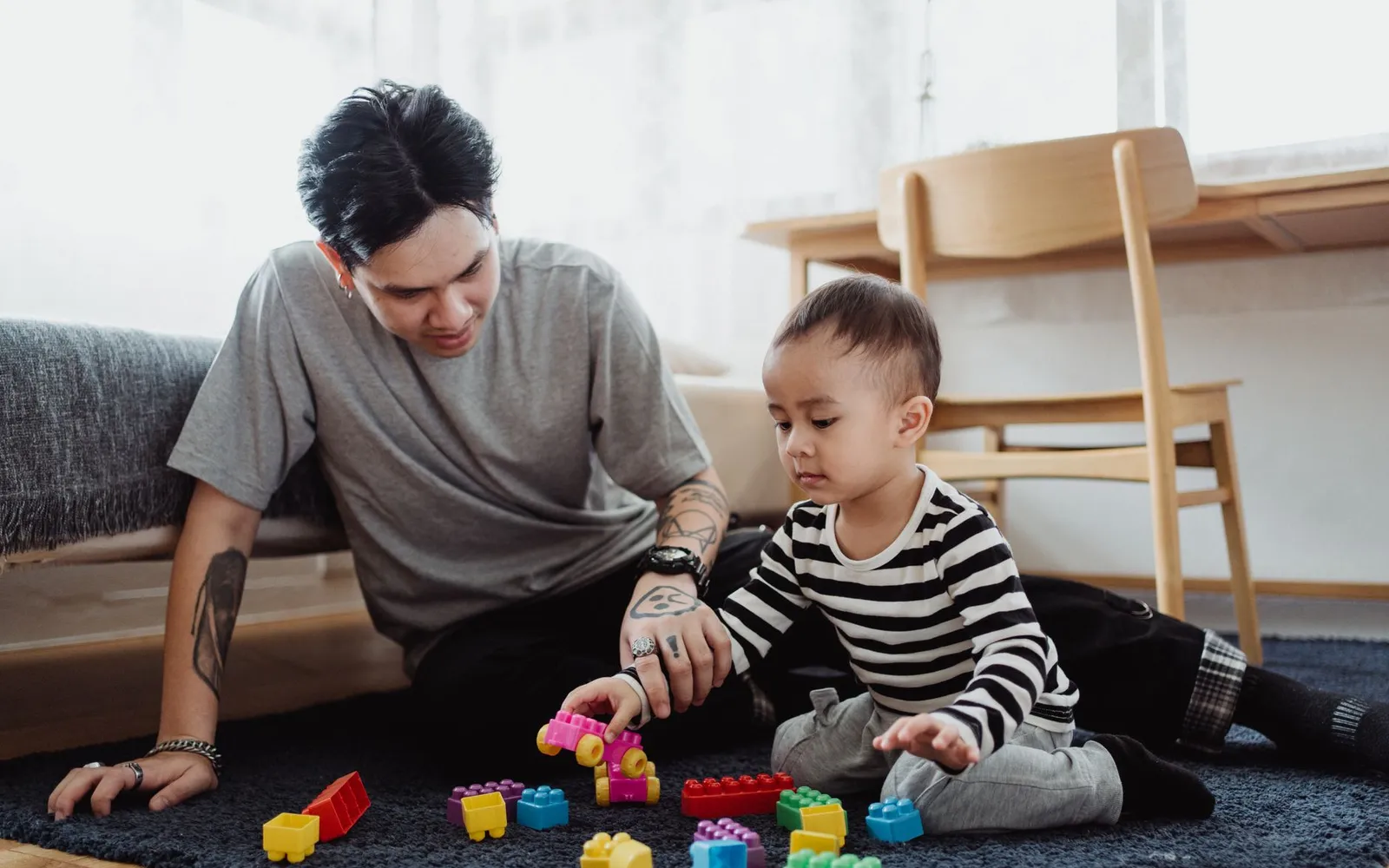 A father sits on a living room rug building with plastic blocks alongside his toddler son