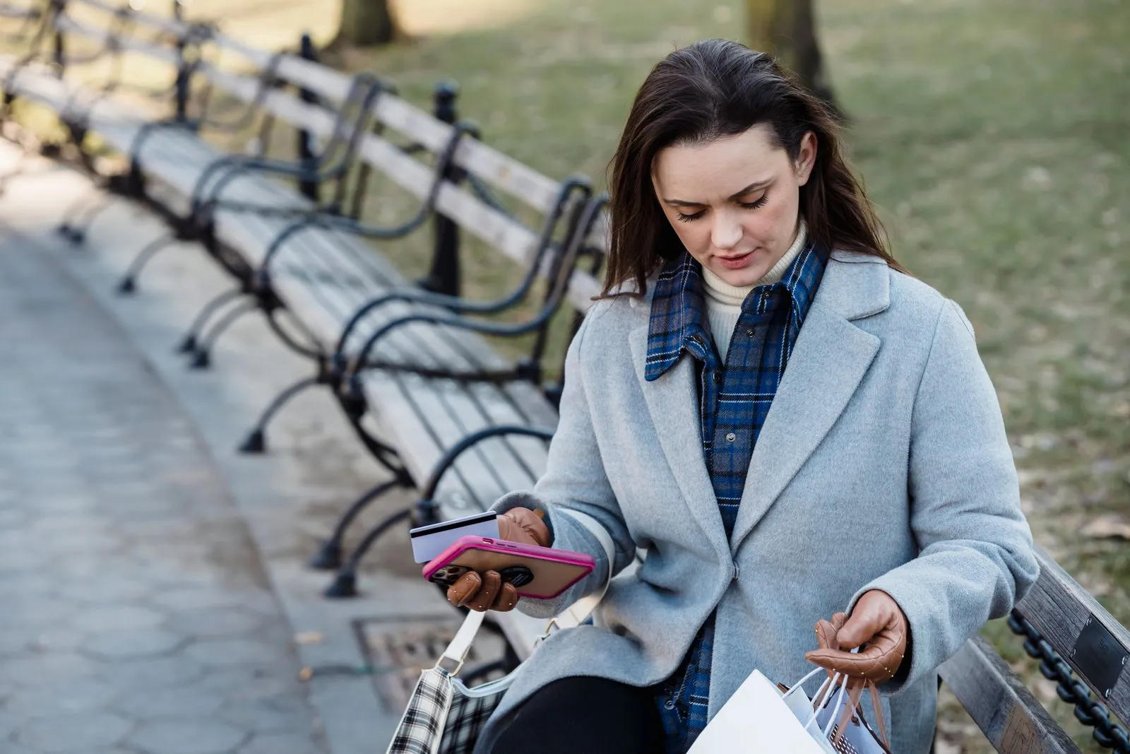 Woman on a park bench holding a phone and a payment card, with shopping bags at her side