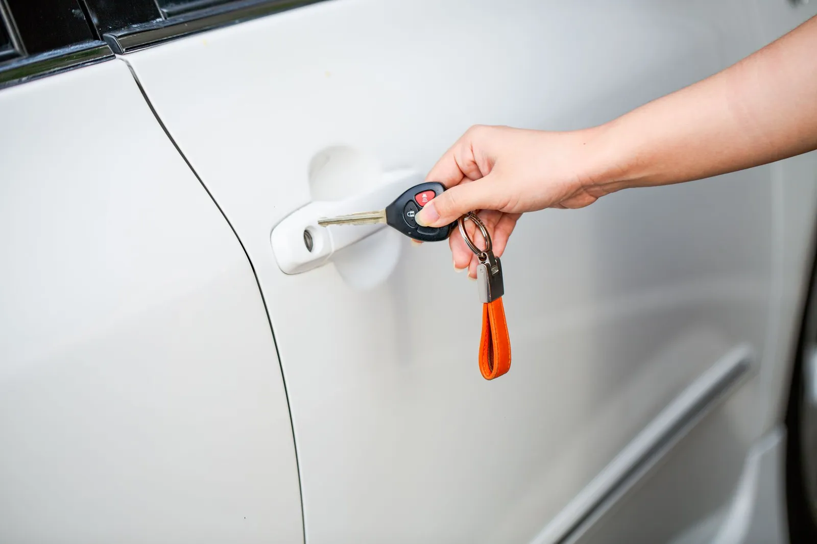 A hand holding a car key and fob beside the door handle of a white car