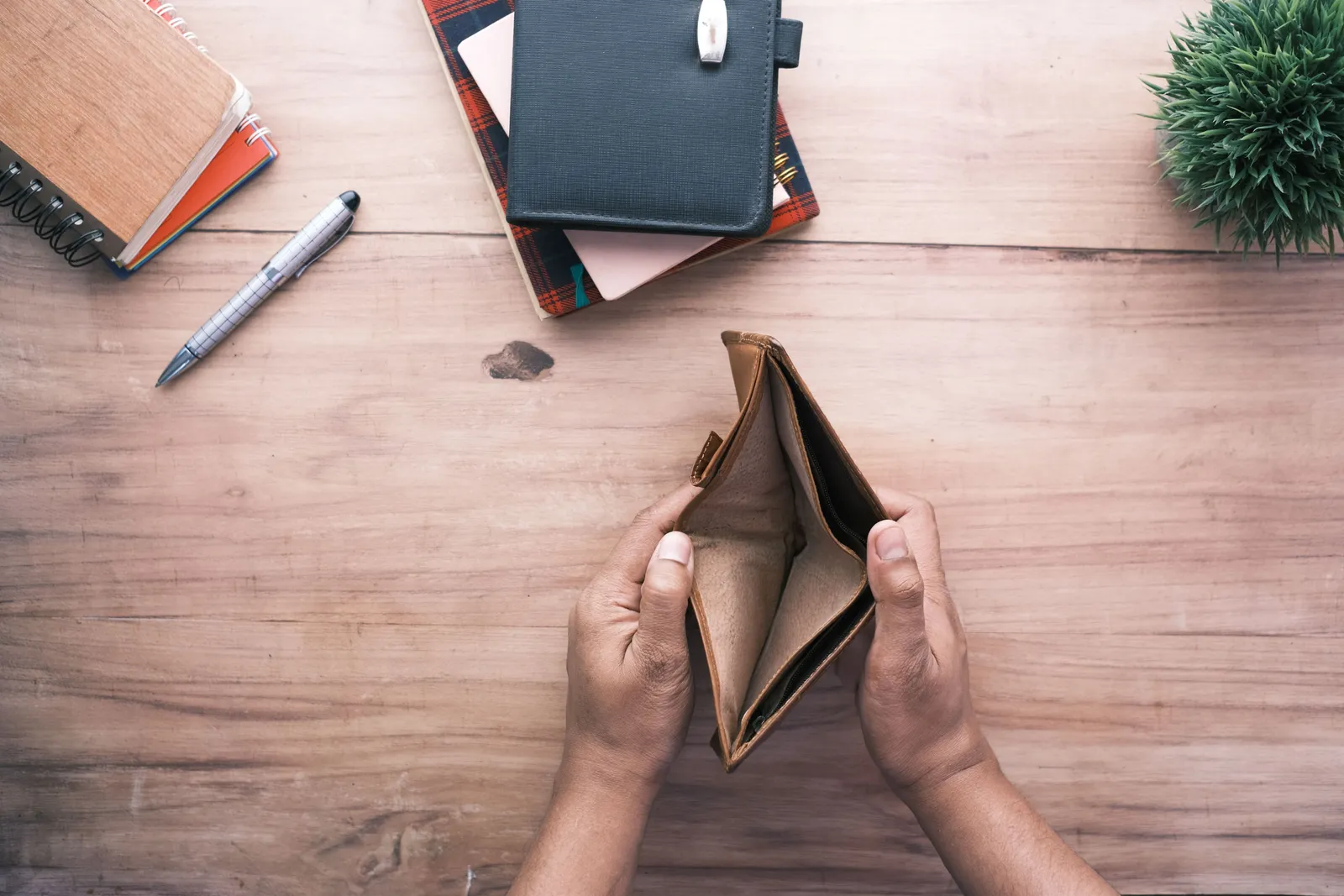 Two hands holding open an empty brown leather wallet over a wooden desk