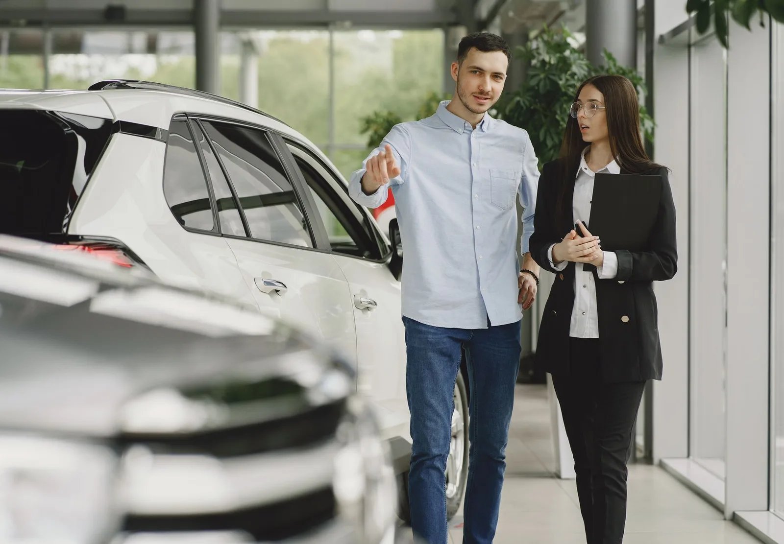 A car shopper points toward a white SUV in a showroom while a salesperson with a folder listens