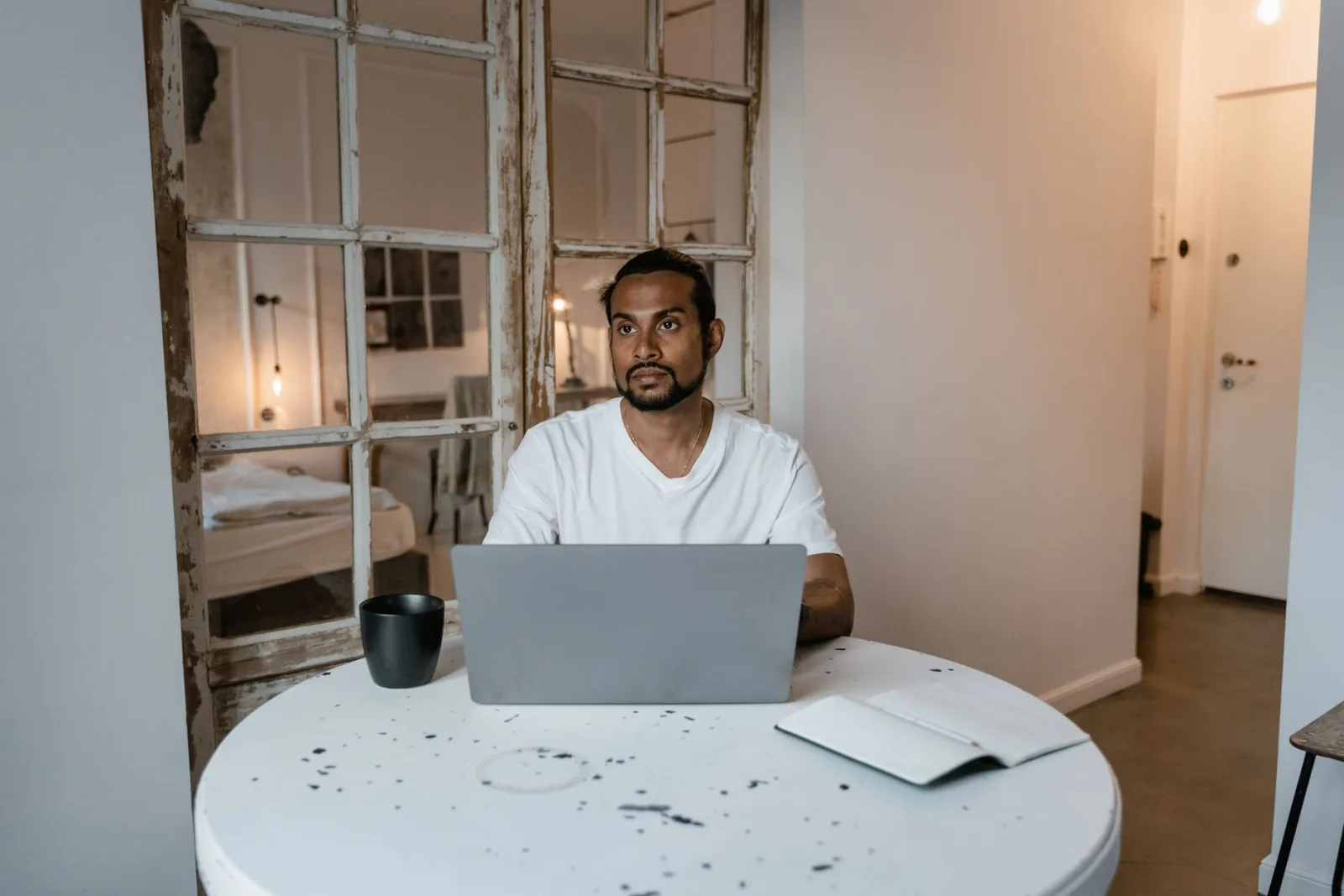 A man sitting at a round kitchen table with a laptop and a coffee cup, thinking