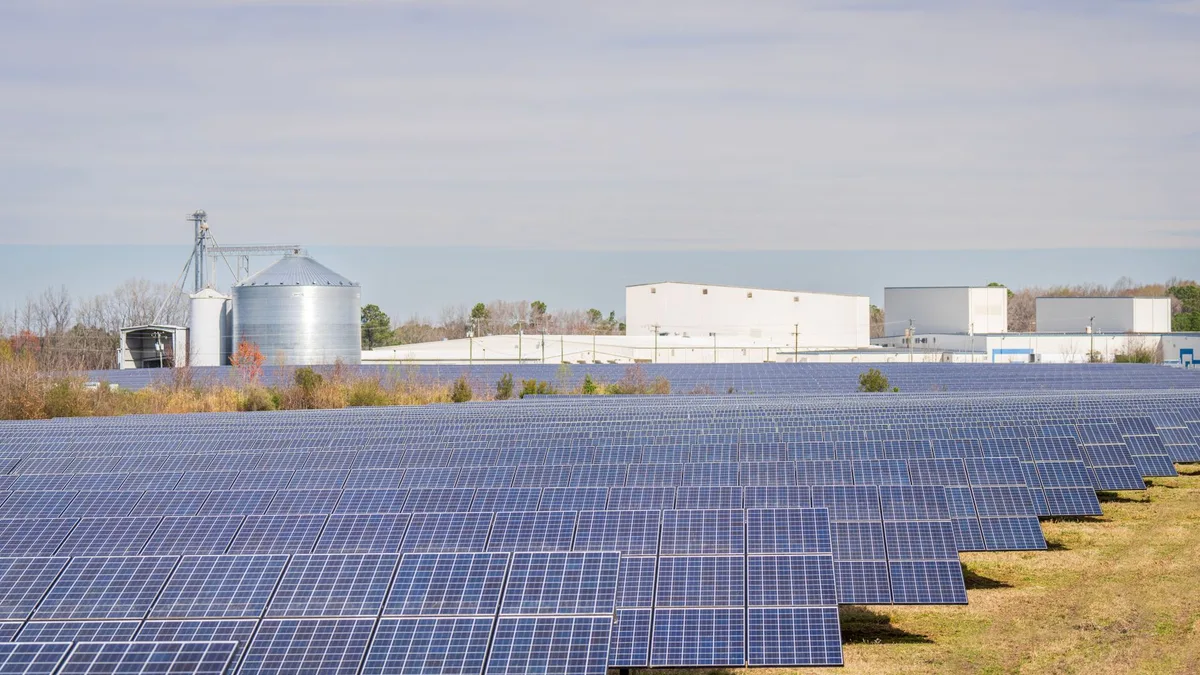 A large field of solar panels with warehouses and silos in the background under a clear sky