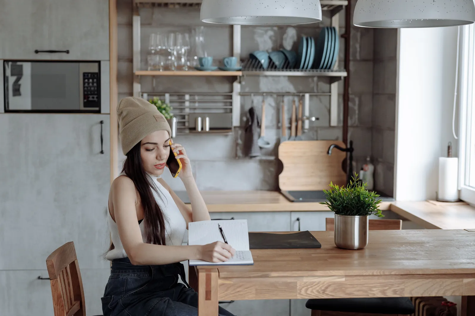 A woman on a phone call at a kitchen table, writing in a notebook