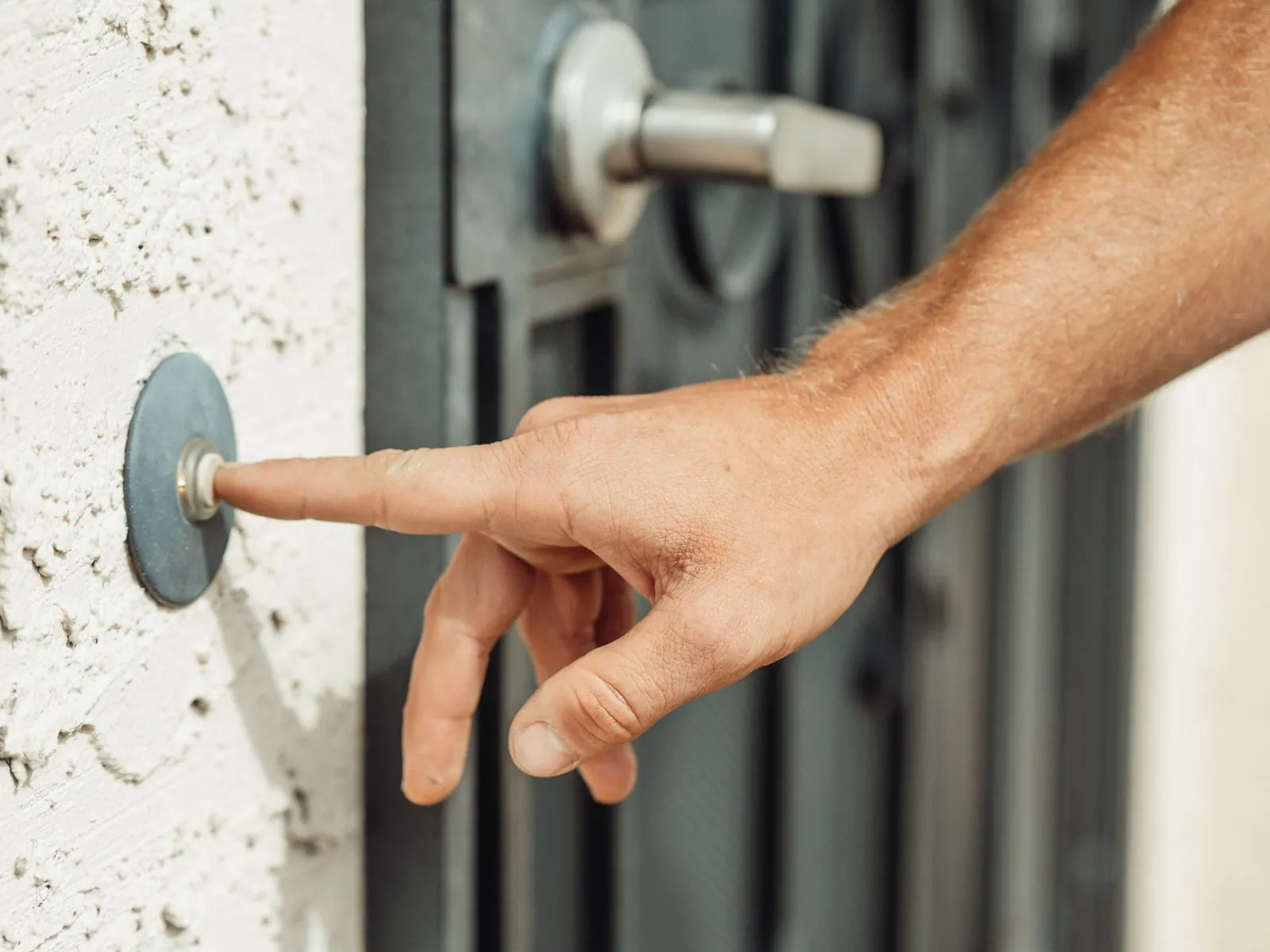 A hand pressing a doorbell on the white wall beside a dark residential door