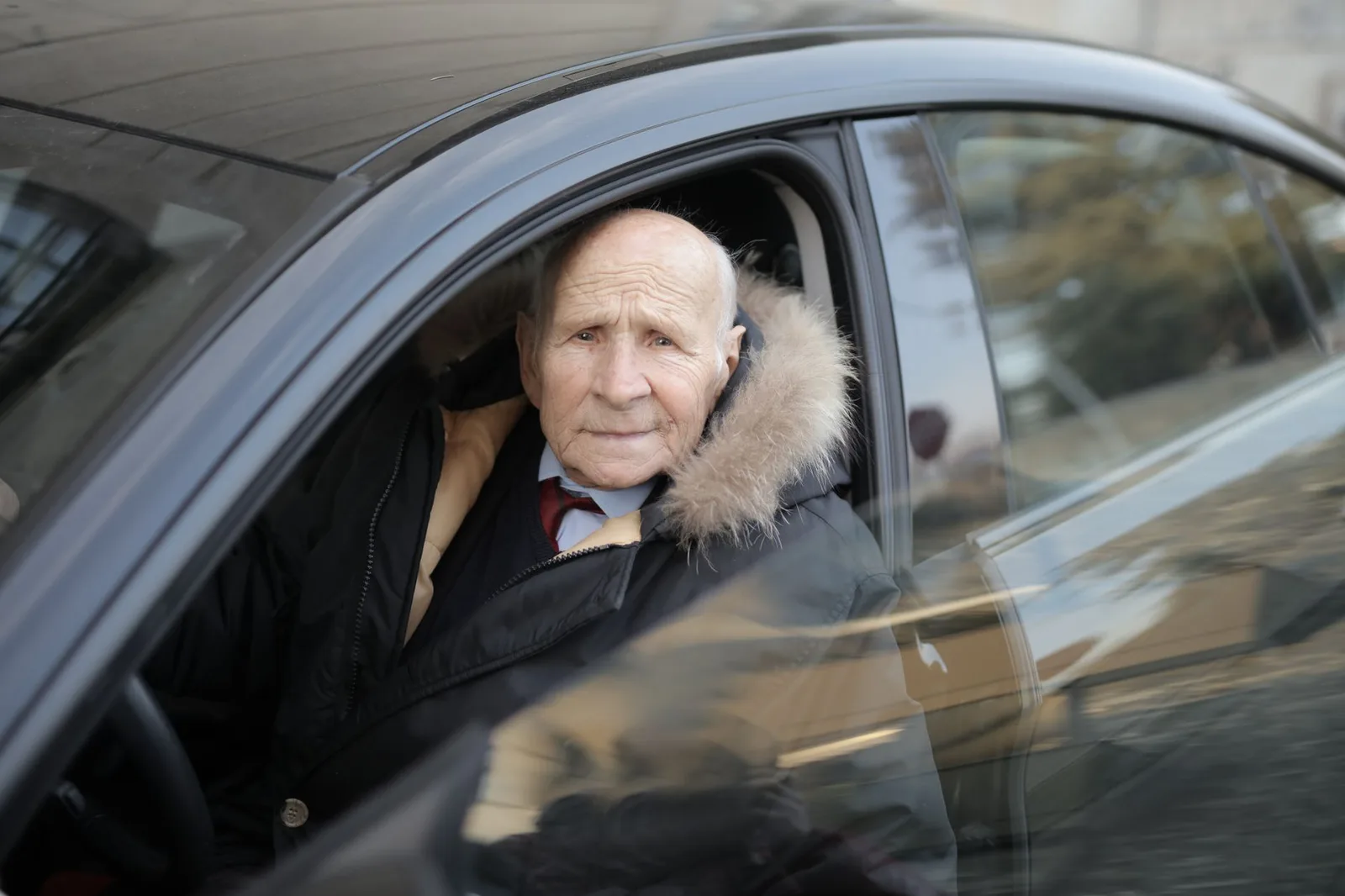 An older man in a winter coat sitting in the driver's seat of a dark car, looking out the open window