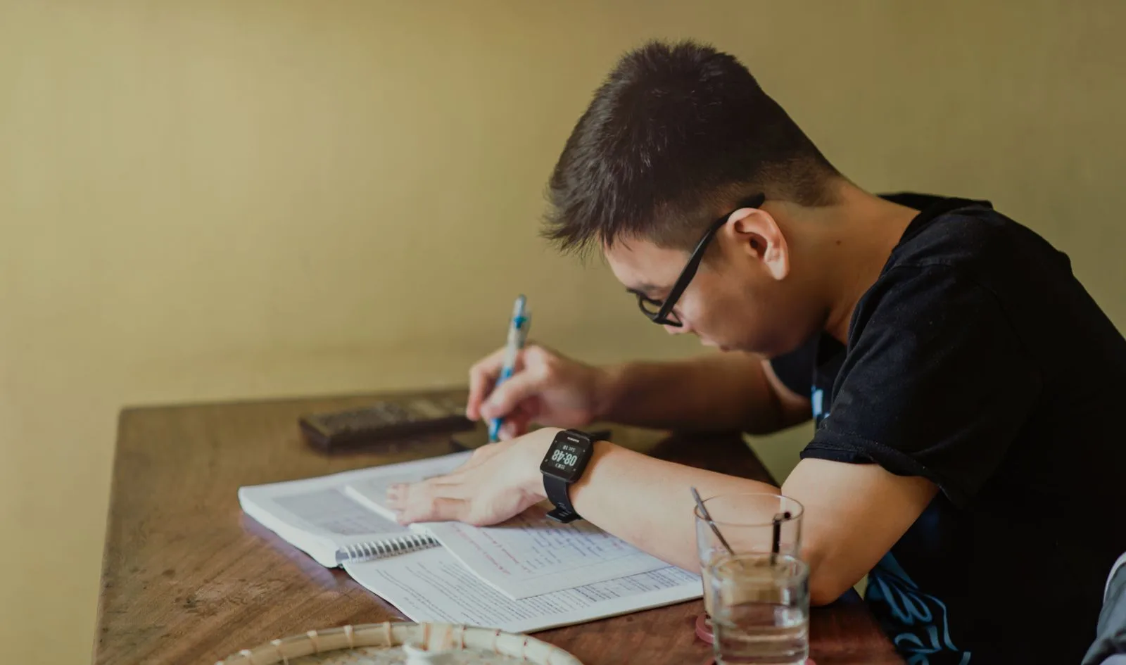 A student in glasses writing notes in a workbook at a wooden table