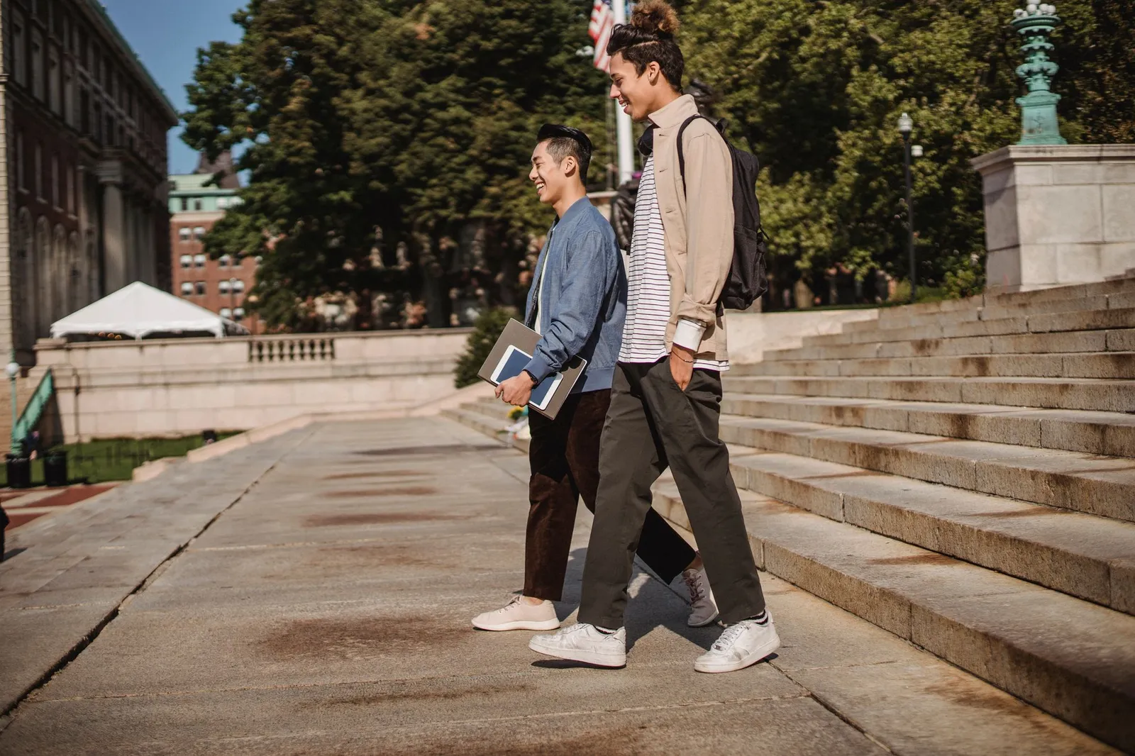 Two college students walking and laughing past stone steps on a sunny campus, one carrying a folder and tablet