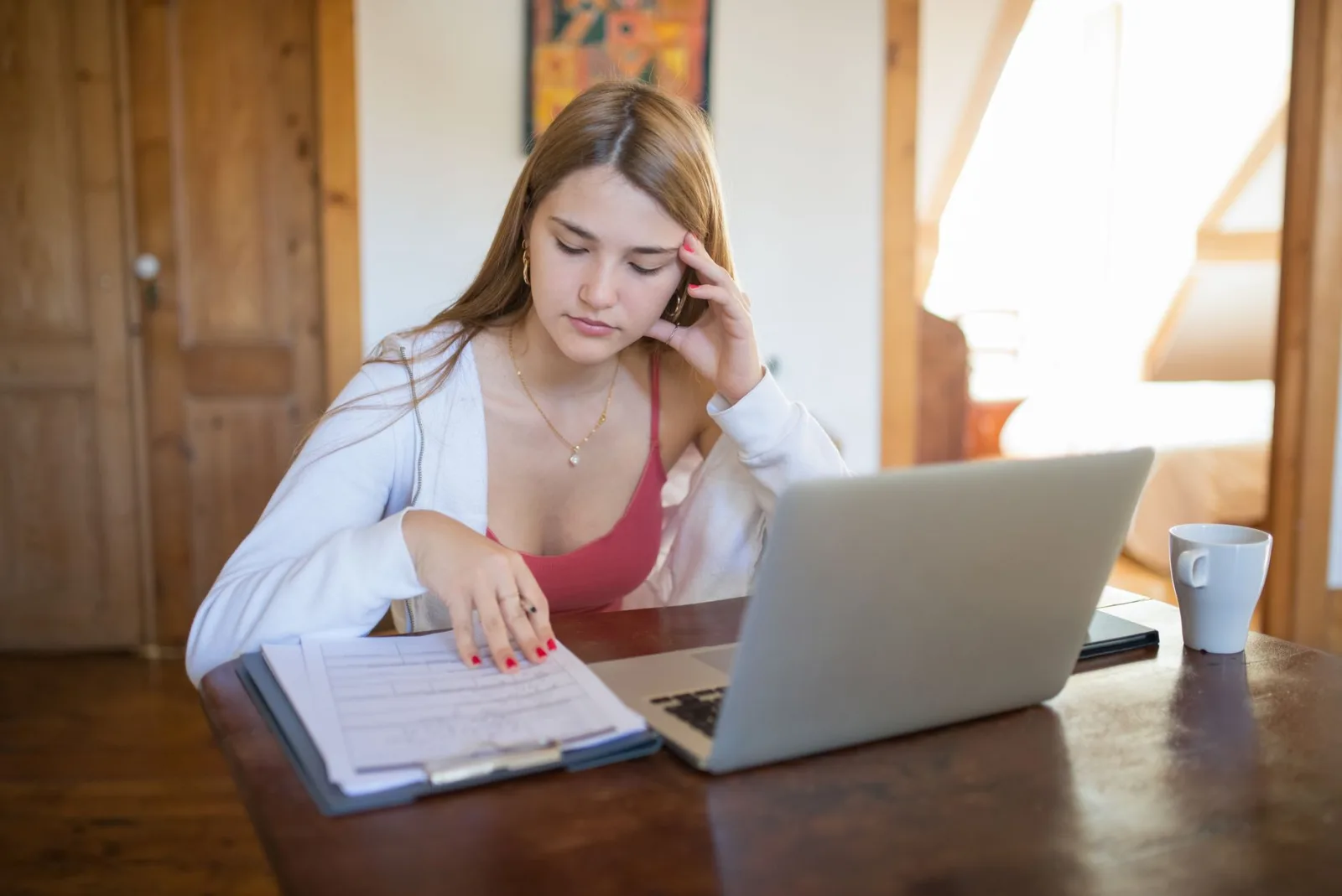Young woman reviewing a clipboard of forms beside a laptop at a kitchen table
