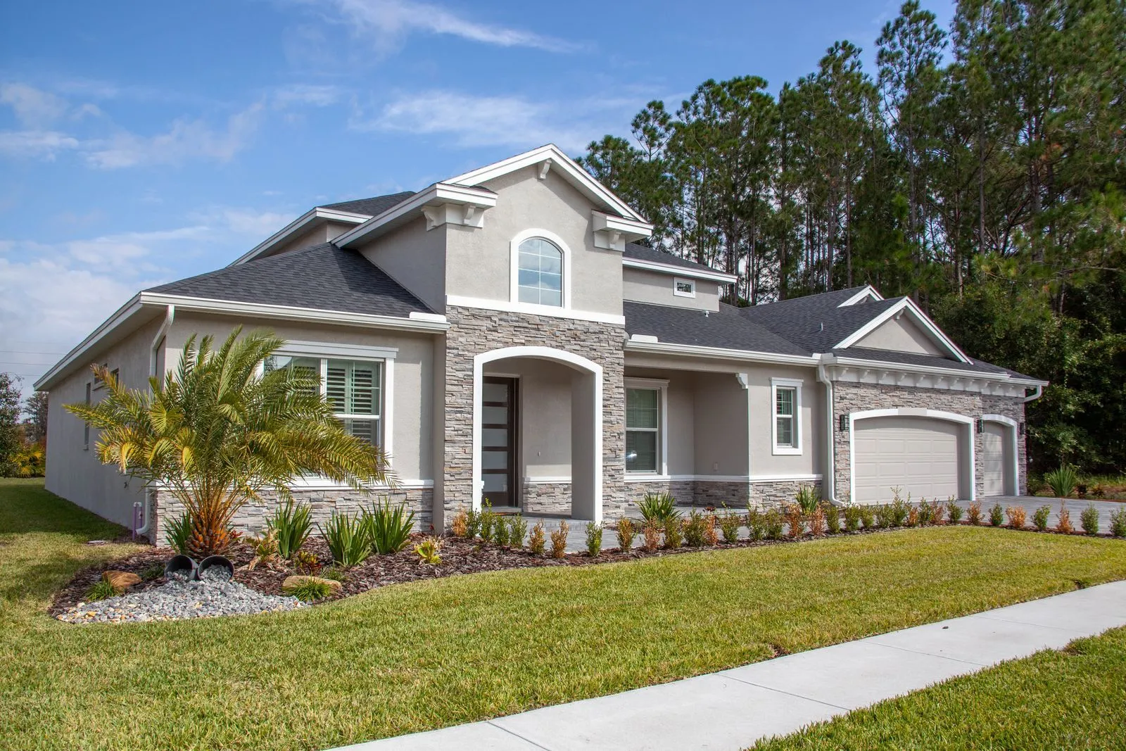 Gray stucco and stone single-story home with a palm tree and a three-car garage under a blue Florida sky
