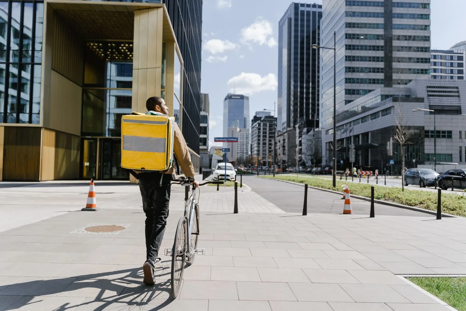 Delivery courier with a plain yellow insulated bag walking a bicycle along a city sidewalk