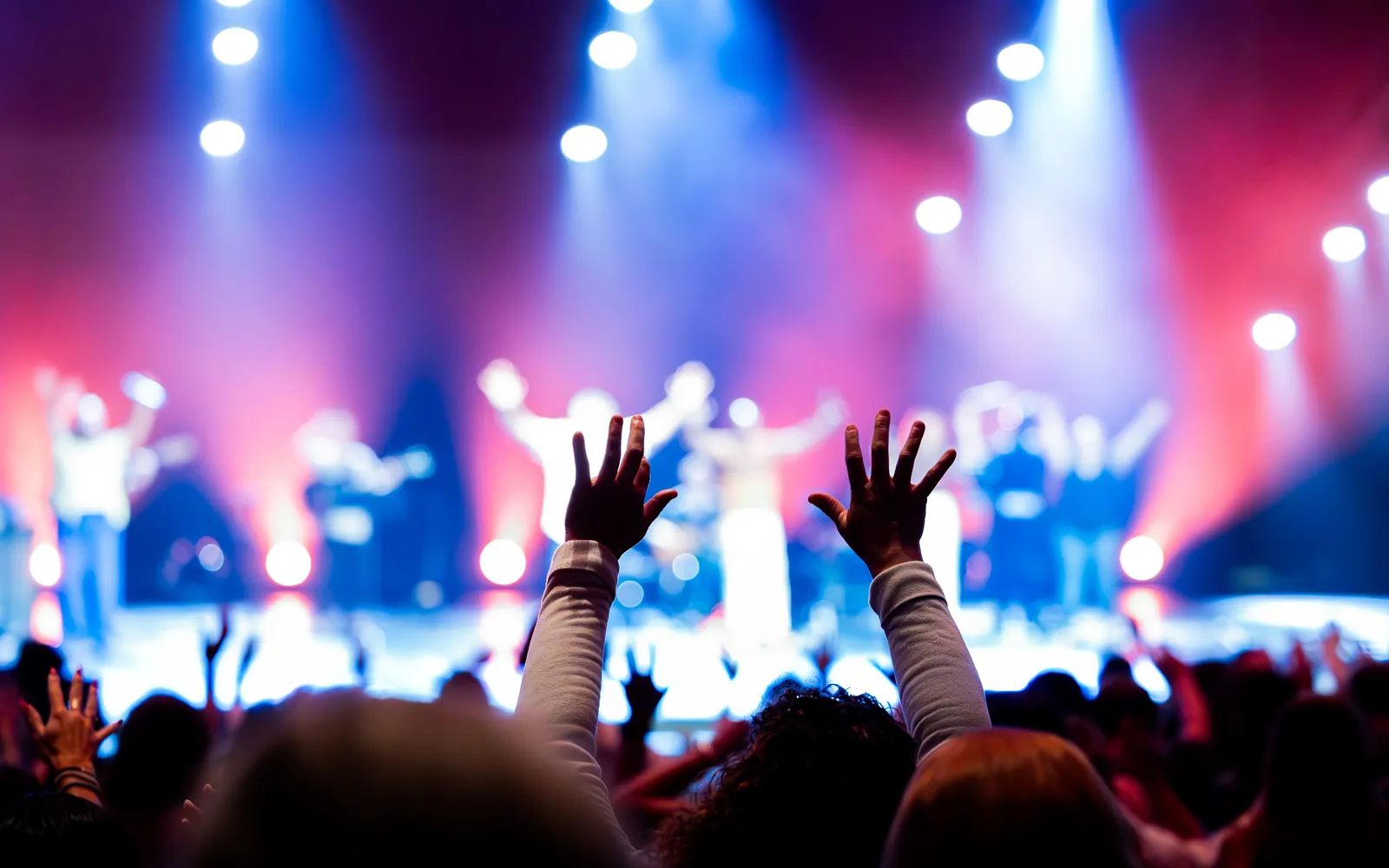 Concert fan seen from behind with both hands raised toward a stage lit in red and blue
