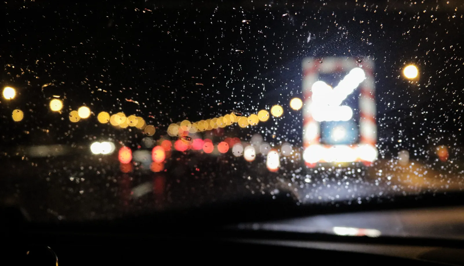 Rain on a car windshield at night, with blurred taillights and a lit road sign ahead