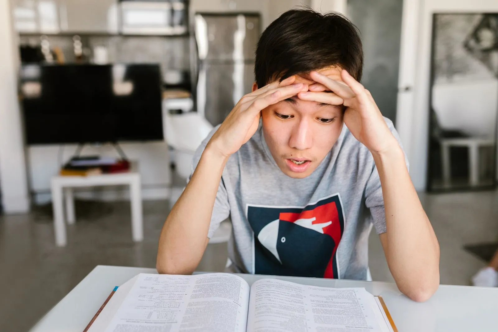A stressed student holding his head over an open textbook at a table in an apartment