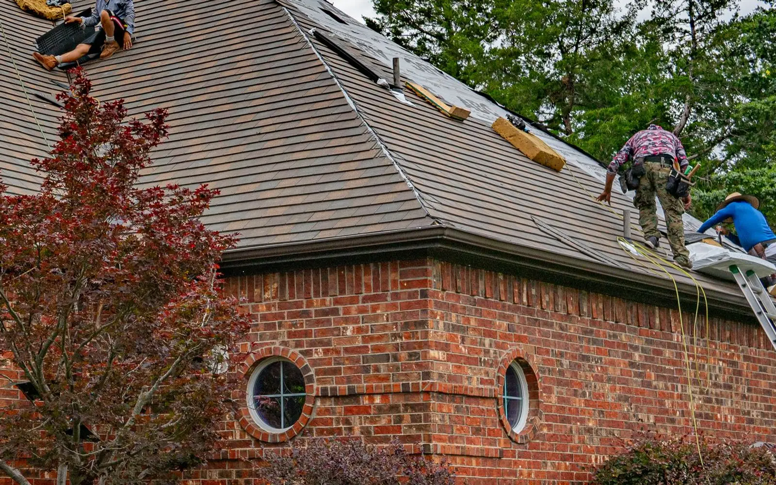 Roofers replacing shingles on a red brick house with a ladder against the gutter
