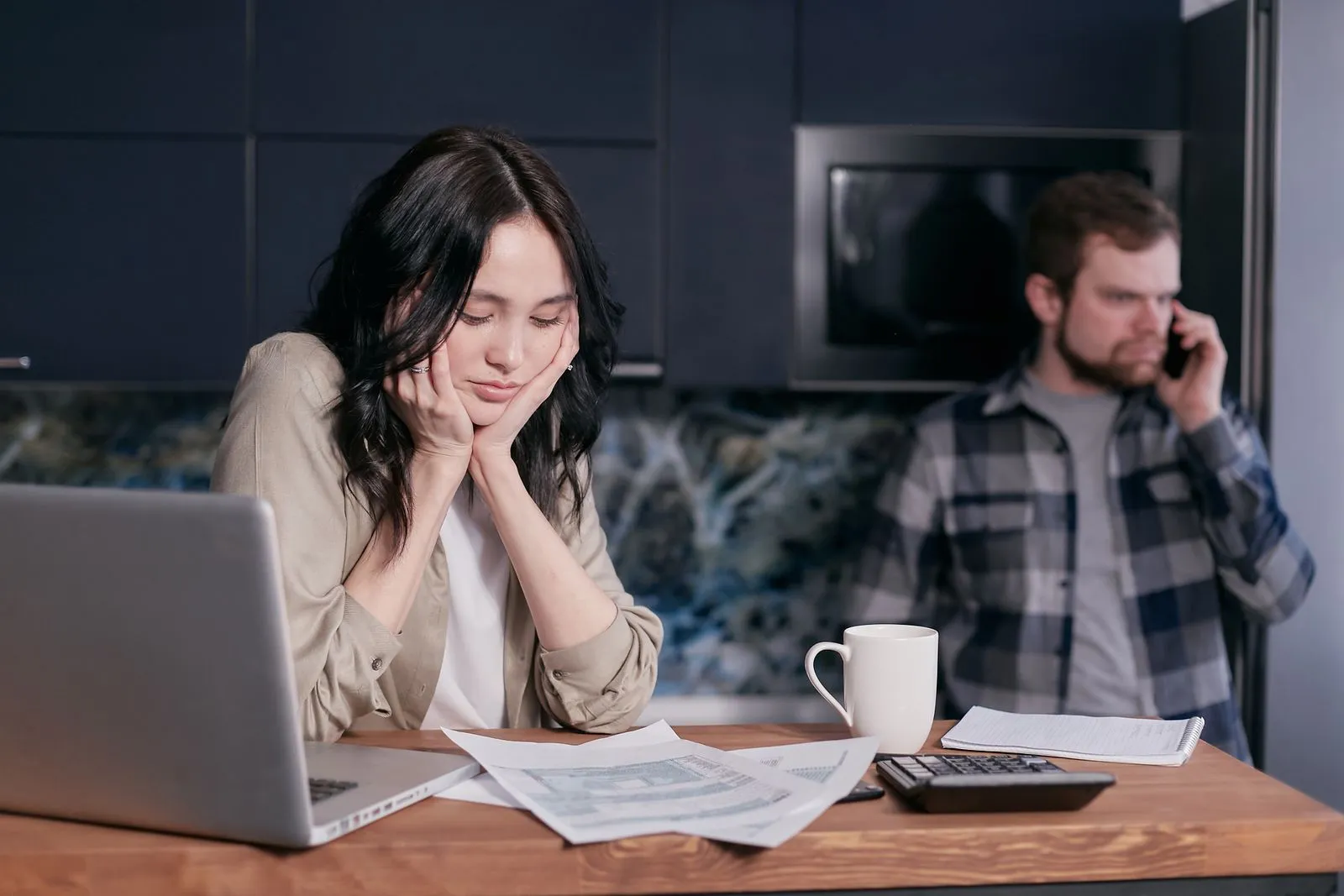 Worried woman at a kitchen table with papers and a laptop while a man behind her talks on the phone