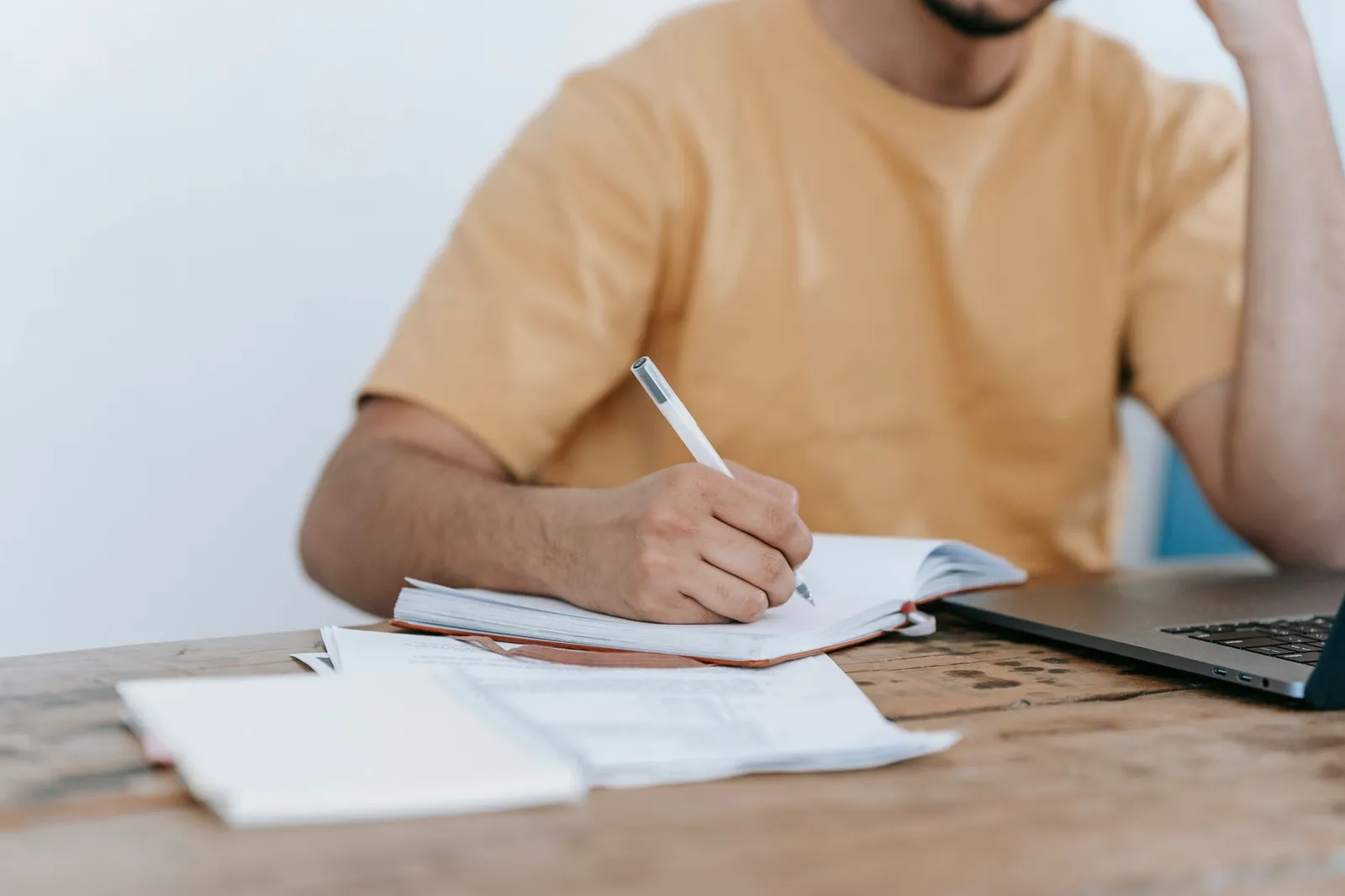 A person writing notes in a notebook beside an open laptop at a wooden table