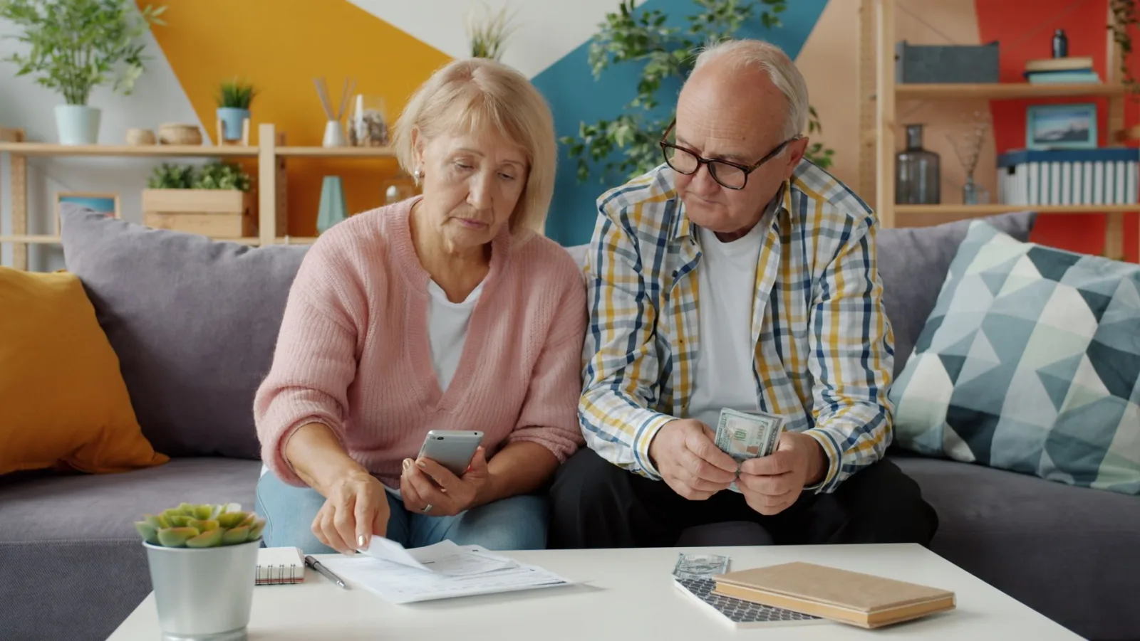 An older couple sitting on a sofa at home reviewing a paper statement with a smartphone and cash on the coffee table