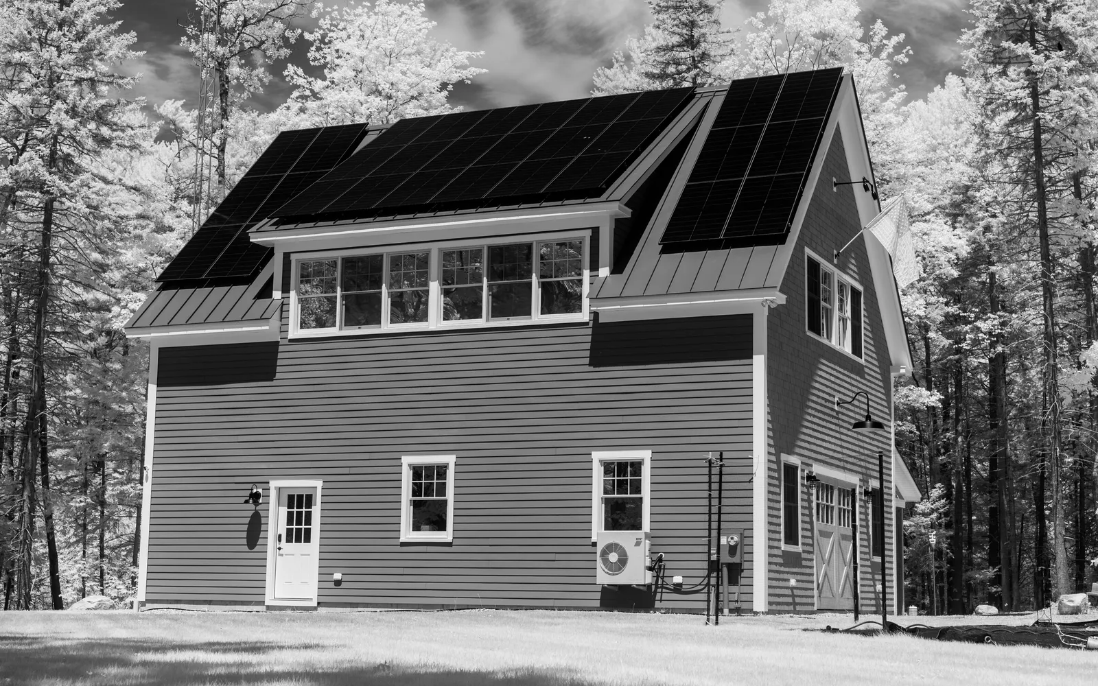 Black and white photo of a clapboard house in the woods with solar panels on its roof and a heat pump on the wall