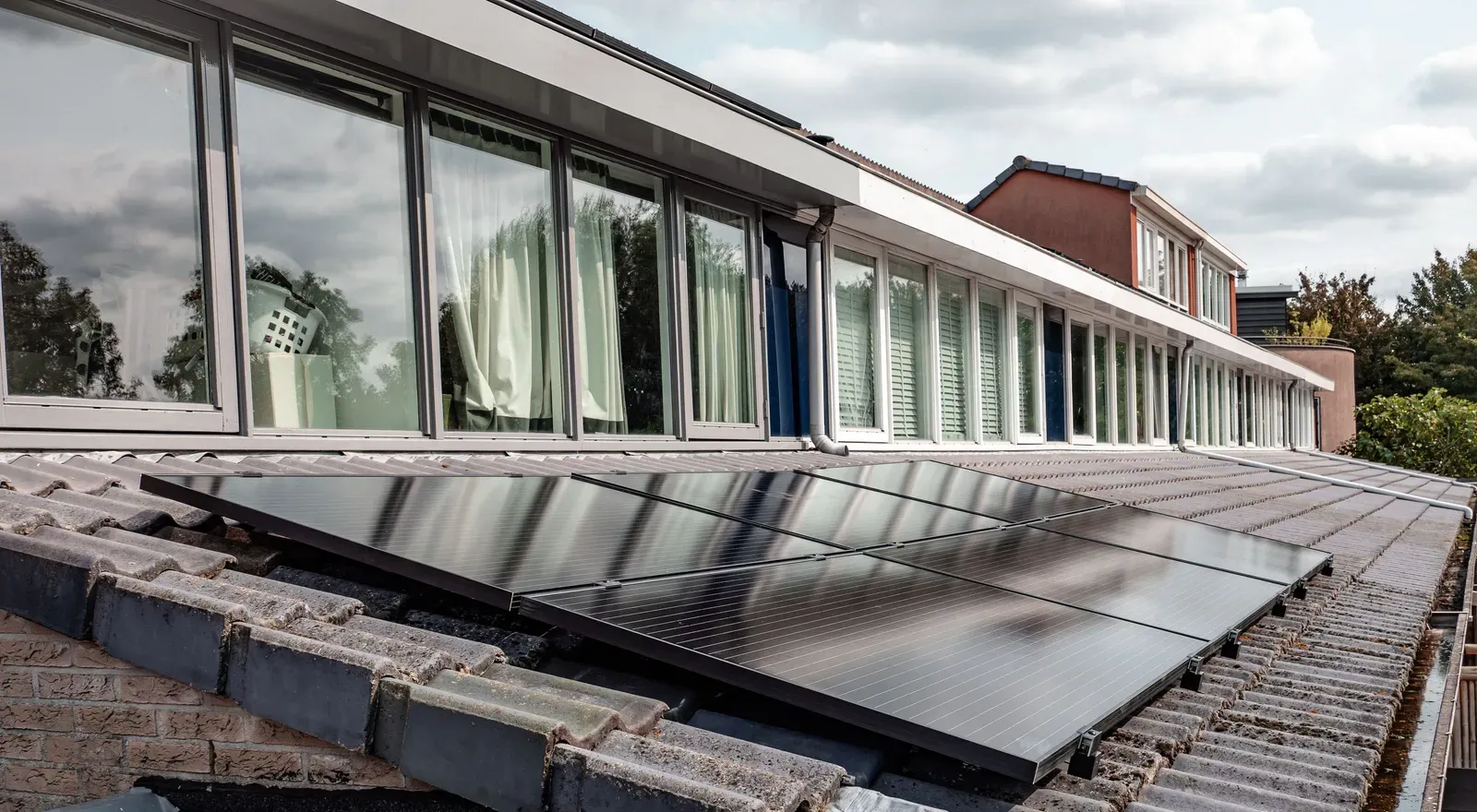 A row of black solar panels mounted on a gray tile roof beneath a line of windows