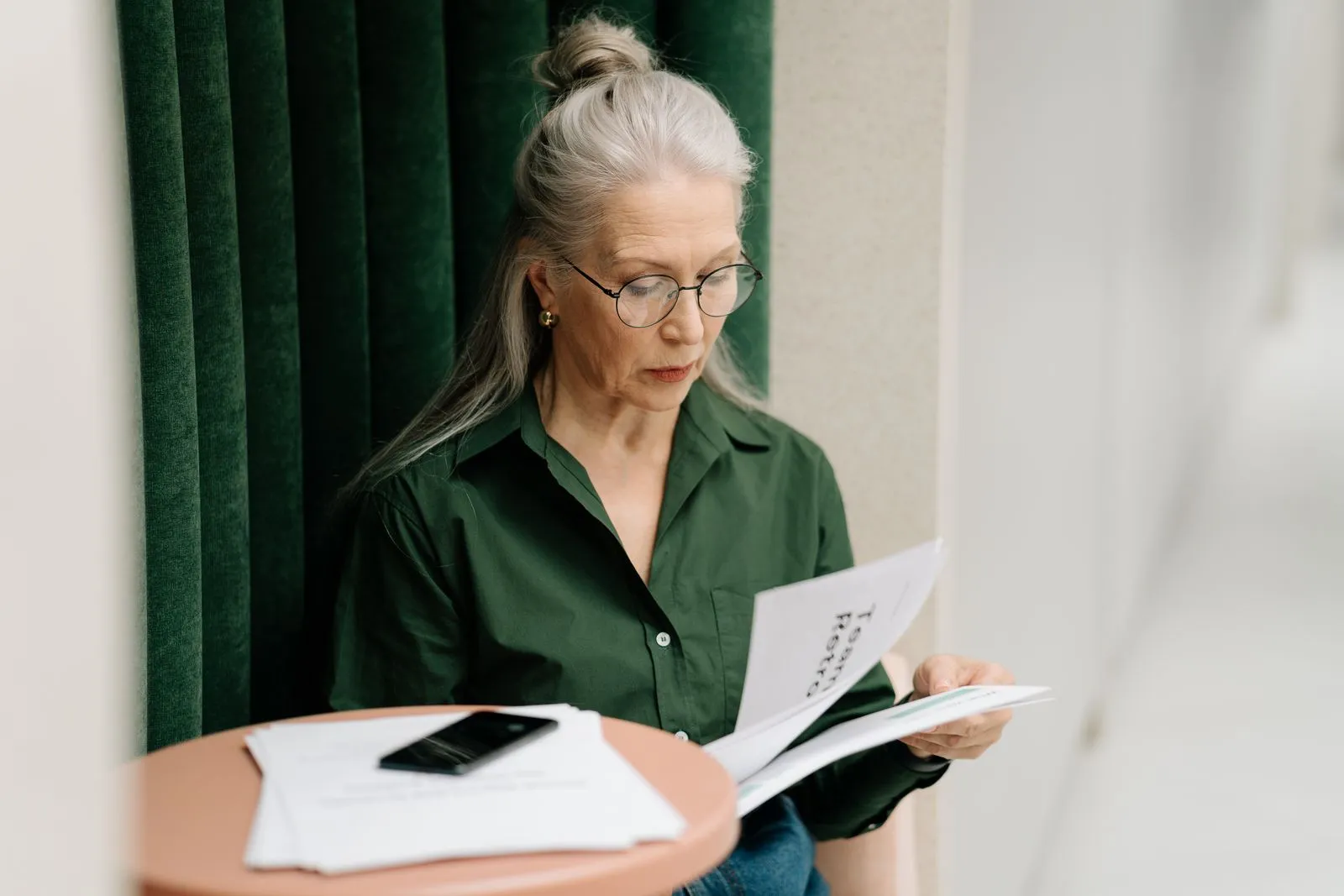 An older woman in glasses reading a stack of printed documents at a small table