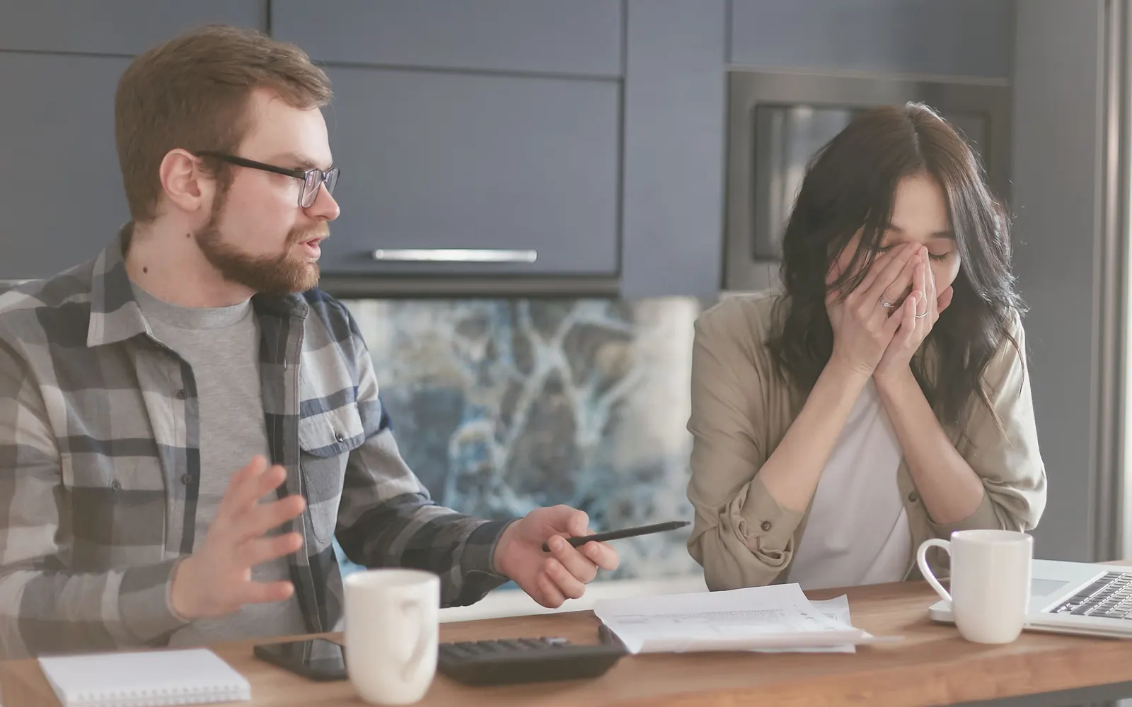 A stressed couple at a kitchen table with papers, a calculator and coffee mugs, one partner covering her face