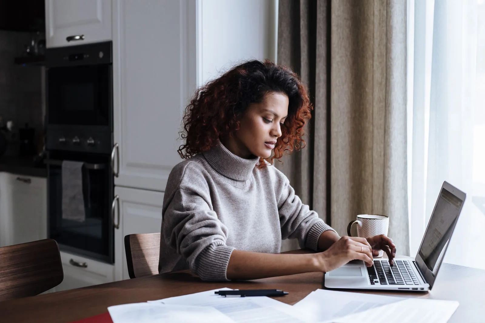 A woman working on a laptop with printed statements spread on a kitchen table