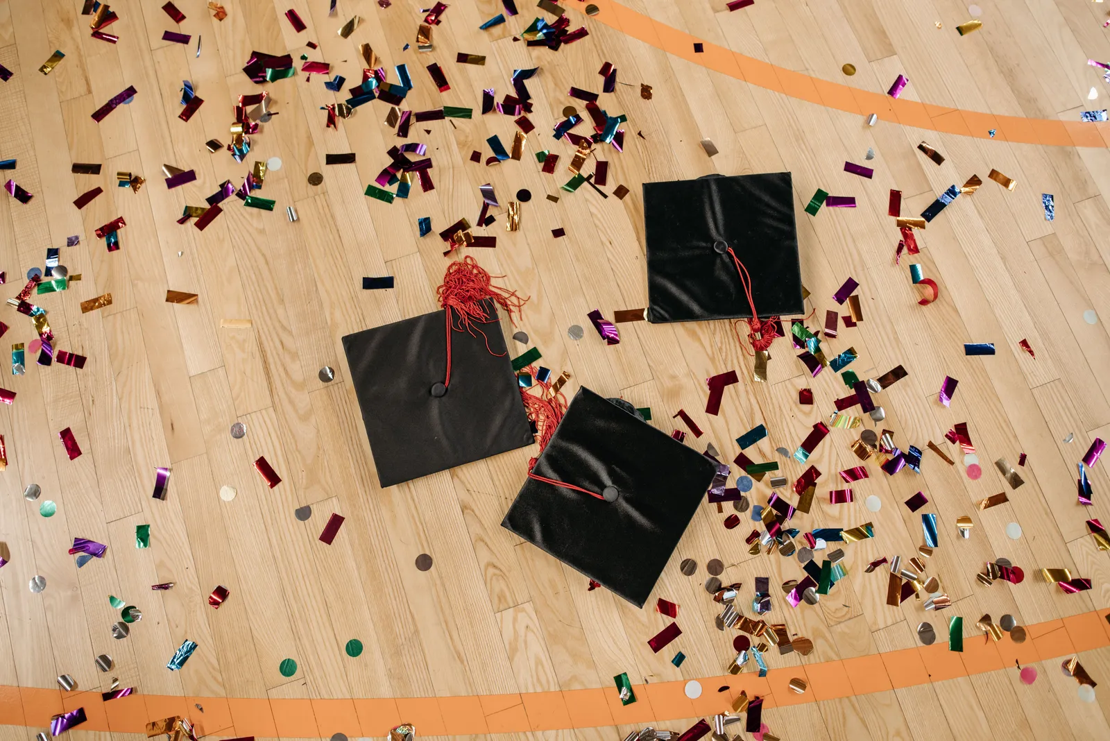 Three black graduation caps with red tassels lying on a gym floor among scattered confetti