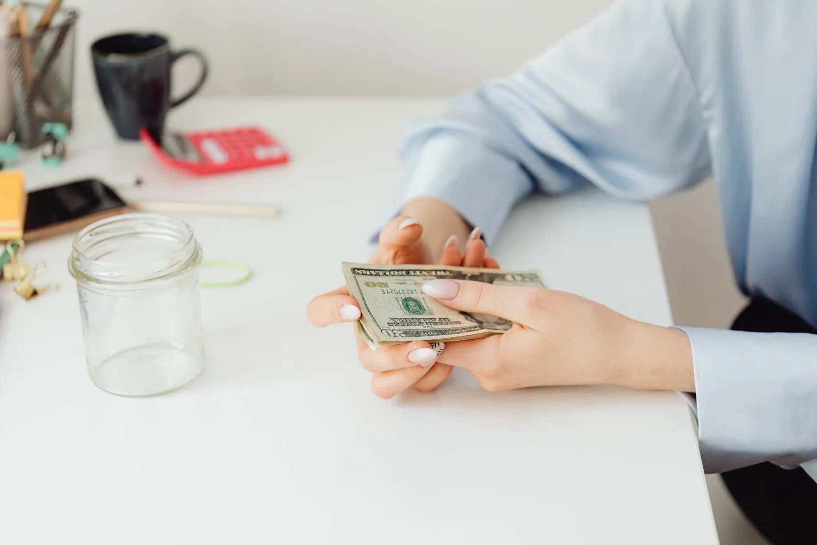 Hands holding a few dollar bills beside an empty glass jar on a white desk