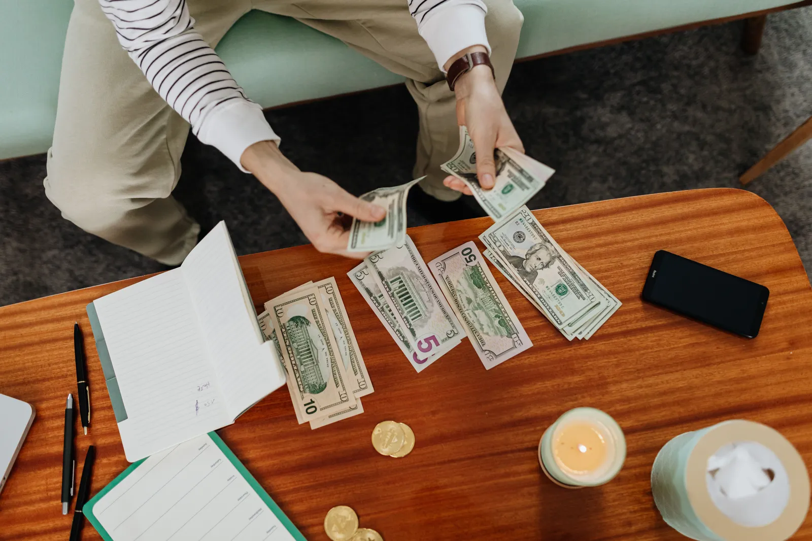 Hands counting US dollar bills on a wooden table beside an open notebook
