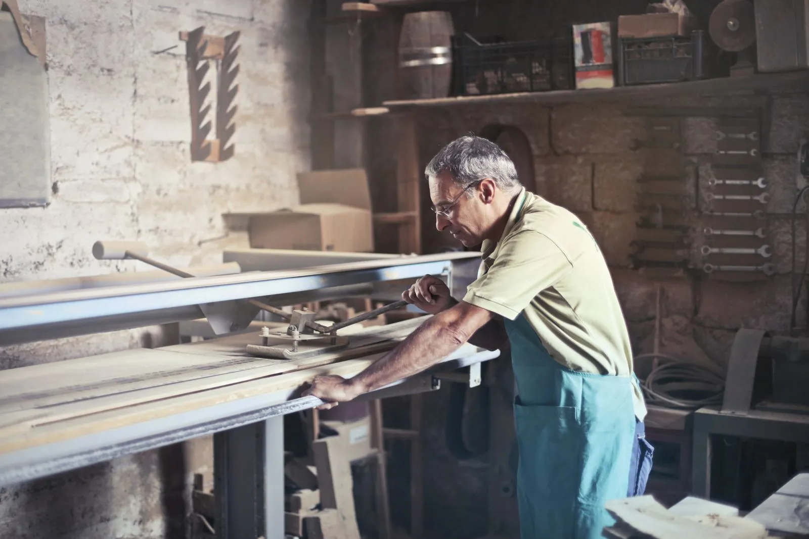 Gray-haired man in glasses and an apron working at a table saw in a stone-walled workshop