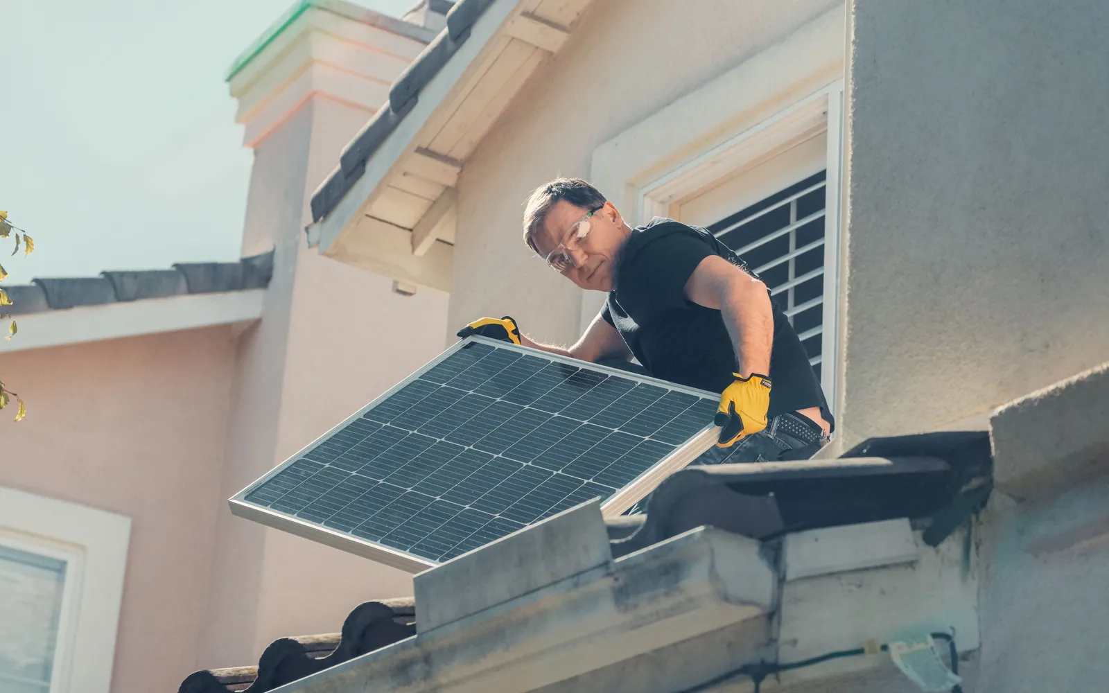 Installer in safety glasses and work gloves lifting a solar panel onto a tiled house roof