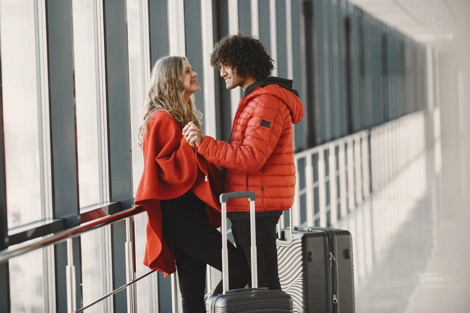 Couple holding hands by the windows of an airport walkway with two rolling suitcases