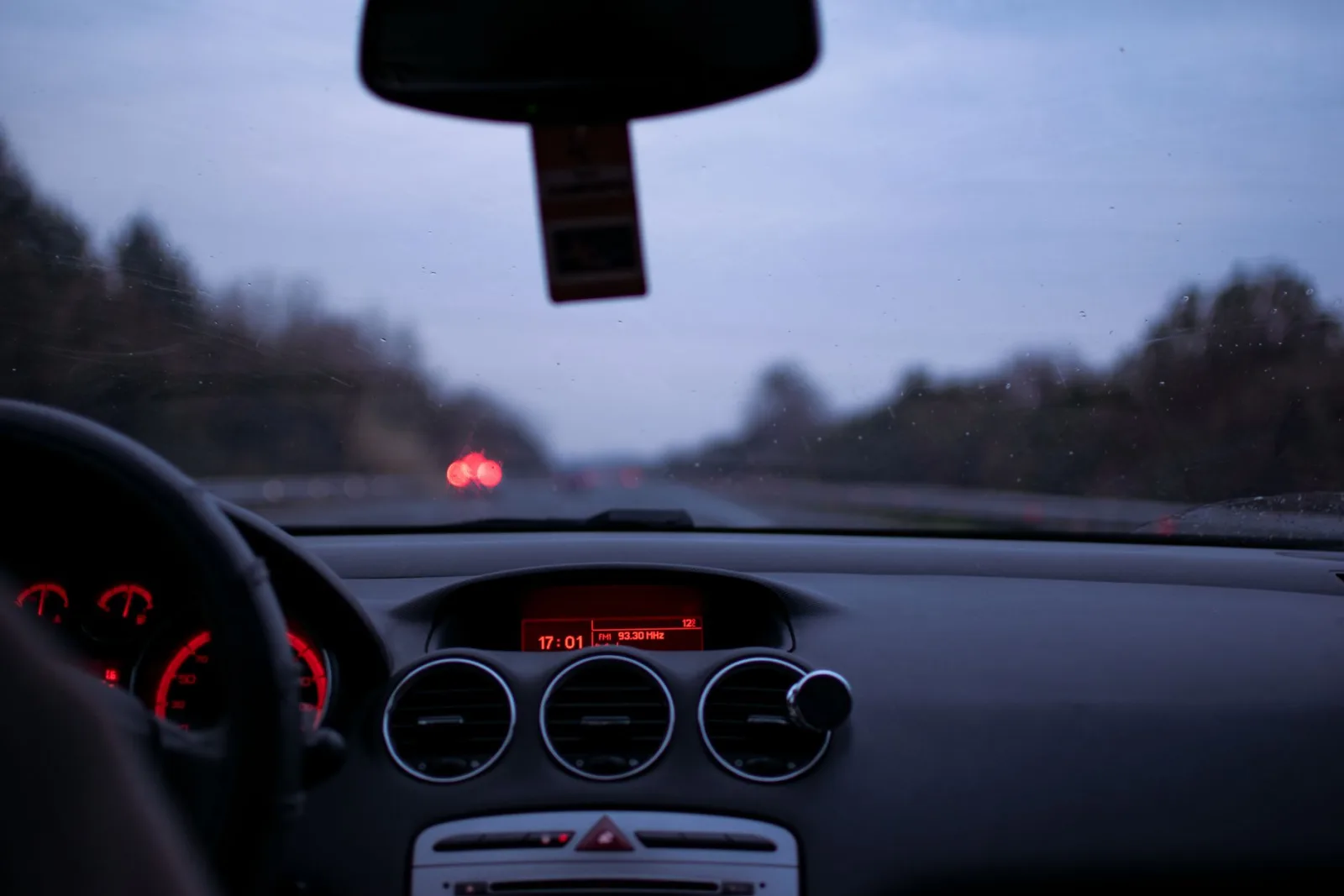 A driver's view through a car windshield at dusk with the dashboard lit in red